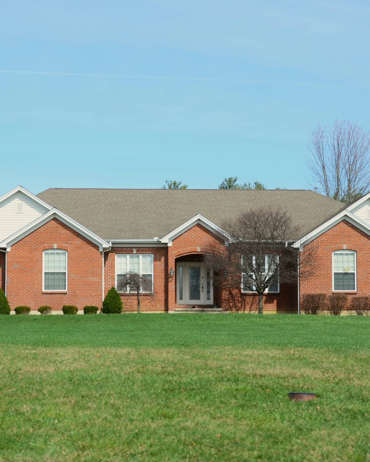 Single-story red brick ranch home with an attached garage, a wide green lawn and a clear blue sky